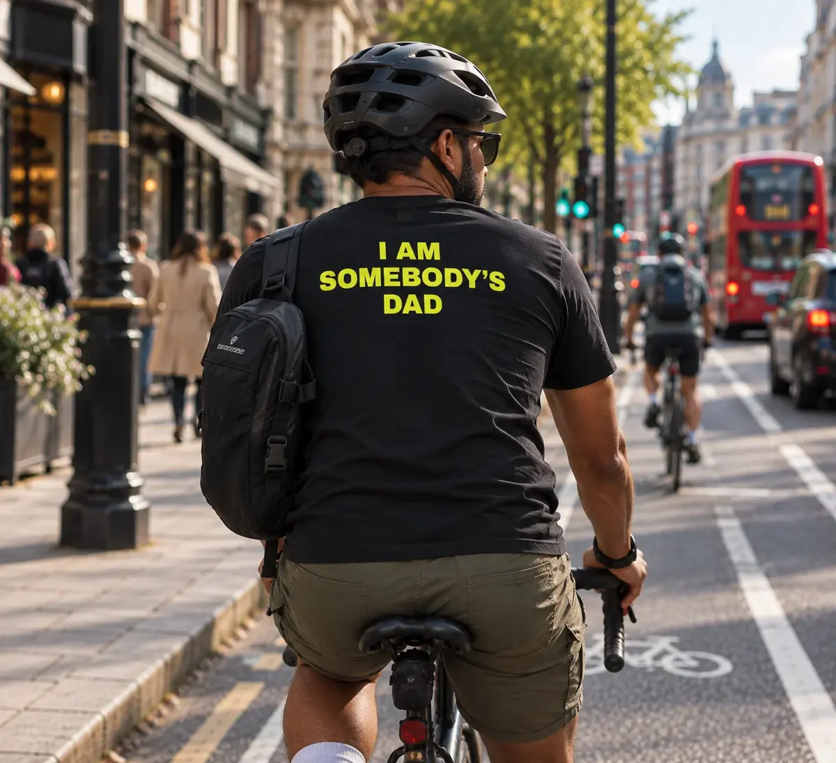 A man with a backpack cycling in a London bike lane, black t-shirt reading “I am somebody’s dad” in yellow